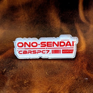 May include: A clear acrylic pin with the text "ONO-SENDAI" in red and "CBRSPC7" in black. The pin also has a barcode and the text "ロックダウン" in Japanese.