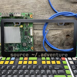 May include: Black license plate frame with the text "source ~/.adventure" in white. The frame is set against a background of computer components, a blue cable, and a colorful keyboard.