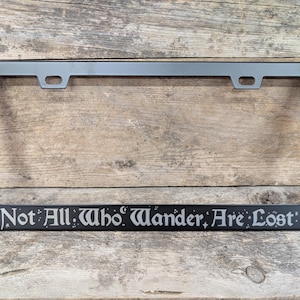May include: Black metal license plate frame with the quote "Not All Who Wander Are Lost" in a decorative font.