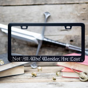 Not all who wander are lost fantasy literary quote engraved on a black stainless steel license plate frame on a woodgrain background of swords, books and dice