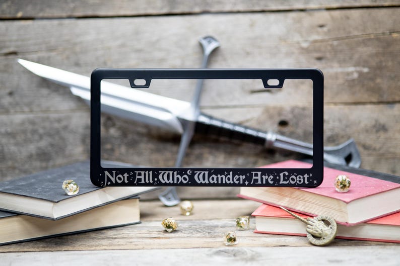 Not all who wander are lost fantasy literary quote engraved on a black stainless steel license plate frame on a woodgrain background of swords, books and dice