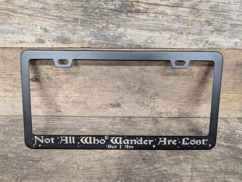 May include: Black metal license plate frame with the text "Not All Who Wander Are Lost." - But I Am.