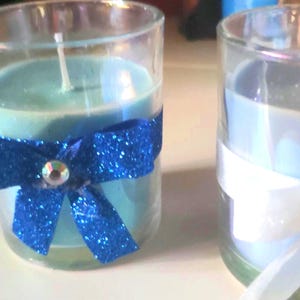 May include: Two glass candle jars with blue and white wax. The jar on the left has a blue bow with a silver rhinestone in the centre. The jar on the right has a white bow with a silver rhinestone in the centre.