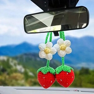 Op de afbeelding: Twee gehaakte aardbei-luchtverfrissers voor de auto met witte bloemen die aan de achteruitkijkspiegel van een auto hangen. De aardbeien zijn rood en groen, en de bloemen zijn wit met gele centra.