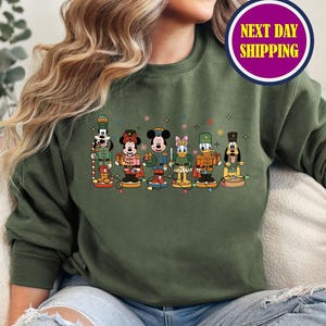May include: Olive green sweatshirt featuring a colorful graphic of Disney characters as nutcrackers. The characters include Mickey Mouse, Donald Duck, and others. The sweatshirt has long sleeves and a crew neck. A circular sticker with the text "NEXT DAY SHIPPING" is visible.