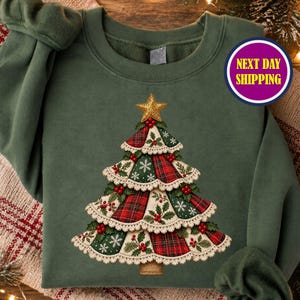 May include: An olive green crewneck sweatshirt featuring a central graphic of a stylized Christmas tree. The tree is composed of layered fabric panels in red plaid, green floral, and snowflake patterns, trimmed with cream lace and red berries.