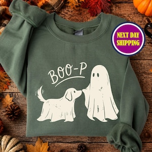 May include: An olive green crewneck sweatshirt featuring a cream-colored graphic of a dog and a ghost facing each other. The text above the figures reads BOO-P in a simple font. The sweatshirt has long sleeves and a ribbed collar and cuffs.