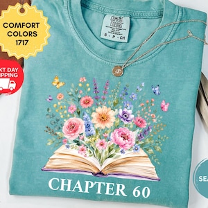 May include: A seafoam green short-sleeve t-shirt featuring a graphic of an open book with a vibrant bouquet of pink, orange, and purple flowers emerging from the pages, accompanied by small butterflies and the text CHAPTER 60 printed below.