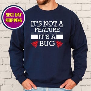 May include: Navy blue sweatshirt with the text "IT'S NOT A FEATURE IT'S A BUG" in white. The design includes two red bug illustrations. A circular graphic with the words "NEXT DAY SHIPPING" is in the upper left corner.