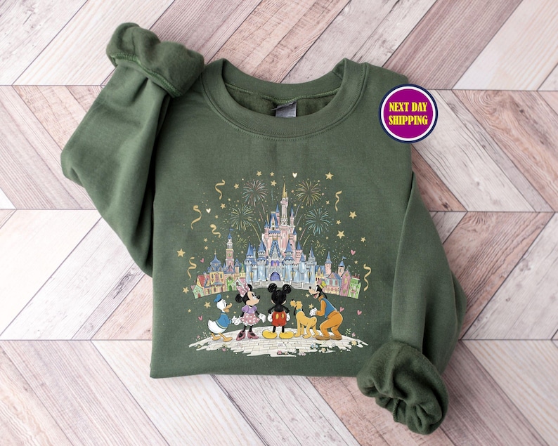 May include: An olive green sweatshirt featuring a colorful graphic of a castle, fireworks, and cartoon characters. The characters include Mickey Mouse, Minnie Mouse, Donald Duck, and Goofy. The sweatshirt has long sleeves and a crew neck.