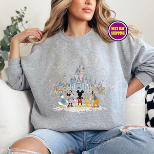 May include: A gray sweatshirt featuring a colorful illustration of a castle and cartoon characters. The design includes Mickey Mouse, Minnie Mouse, Donald Duck, and Goofy. The sweatshirt has long sleeves and a crew neck.