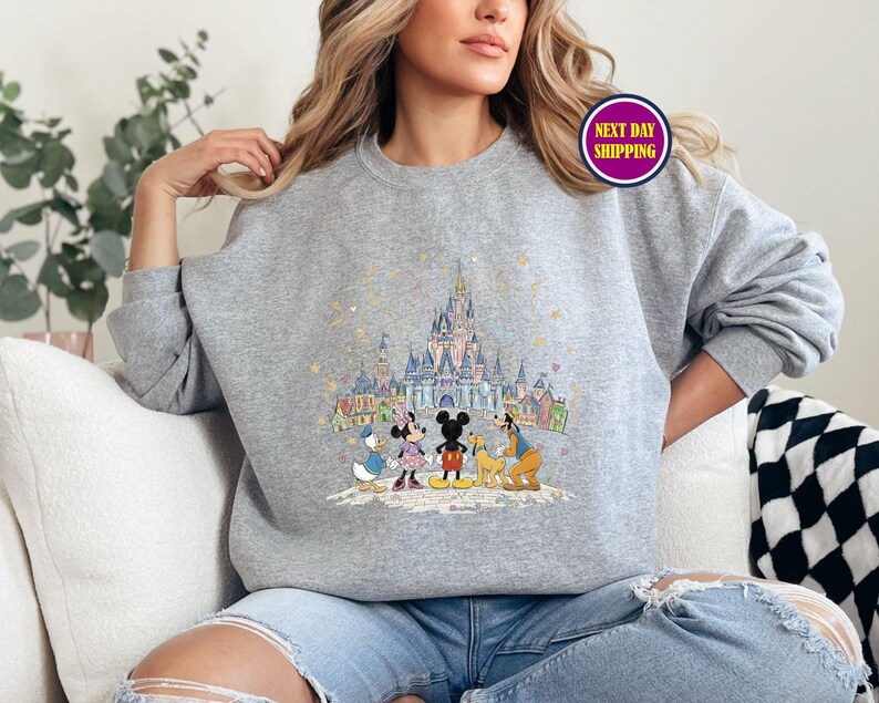 May include: A gray sweatshirt featuring a colorful illustration of a castle and cartoon characters. The design includes Mickey Mouse, Minnie Mouse, Donald Duck, and Goofy. The sweatshirt has long sleeves and a crew neck.
