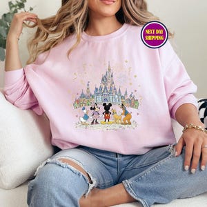 May include: A light pink sweatshirt featuring a colorful illustration of a castle and cartoon characters. The design includes Mickey Mouse, Minnie Mouse, Donald Duck, and Goofy. The sweatshirt has a crew neck and long sleeves.