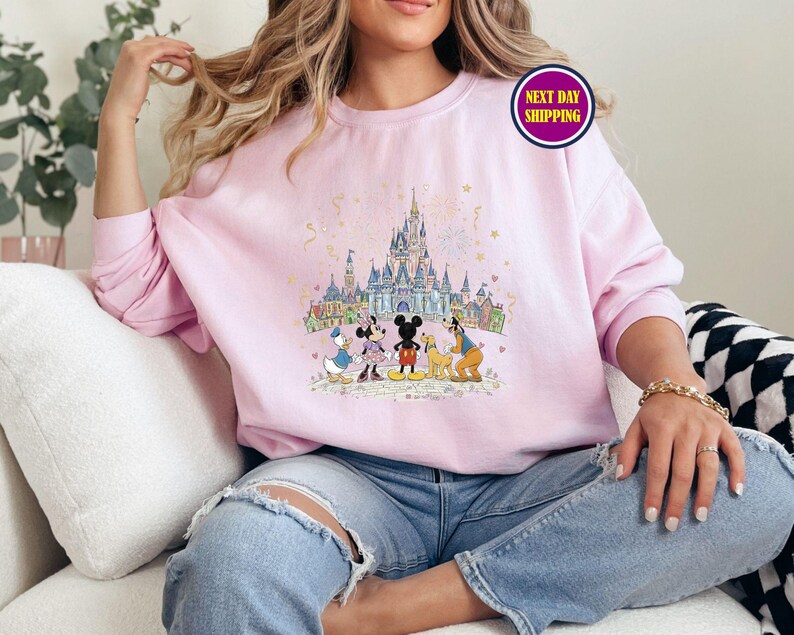 May include: A light pink sweatshirt featuring a colorful illustration of a castle and cartoon characters. The design includes Mickey Mouse, Minnie Mouse, Donald Duck, and Goofy. The sweatshirt has a crew neck and long sleeves.