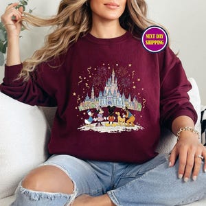 May include: Burgundy crewneck sweatshirt featuring a colorful graphic of a castle, fireworks, and cartoon characters. The design includes Mickey Mouse, Minnie Mouse, Donald Duck, and Goofy. The sweatshirt has long sleeves and a relaxed fit.