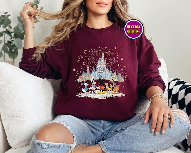 May include: Burgundy crewneck sweatshirt featuring a colorful graphic of a castle, fireworks, and cartoon characters. The design includes Mickey Mouse, Minnie Mouse, Donald Duck, and Goofy. The sweatshirt has long sleeves and a relaxed fit.
