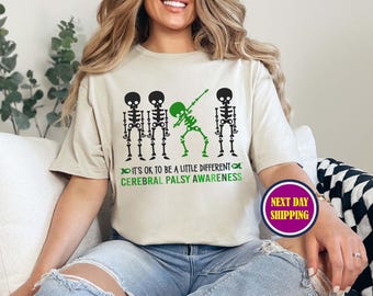May include: A white t-shirt with a green, black, and white skeleton graphic. The skeleton is doing the dab. The text on the shirt reads "It's ok to be a little different Cerebral Palsy Awareness".