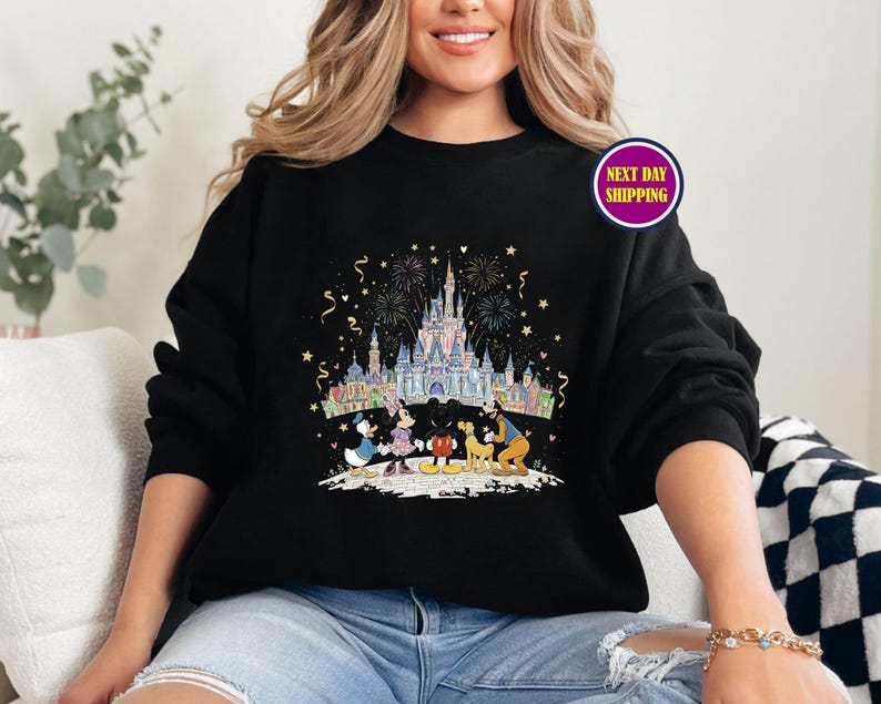 May include: Black sweatshirt featuring a colorful illustration of a castle, fireworks, and cartoon characters. The design includes Donald Duck, Mickey Mouse, and Minnie Mouse. The sweatshirt has a crew neck and long sleeves.