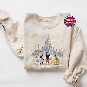 May include: Beige crewneck sweatshirt featuring a colorful illustration of a castle and cartoon characters. The graphic includes Mickey Mouse, Minnie Mouse, Donald Duck, and Goofy. The sweatshirt has the text "Next Day Shipping" on a circular sticker.