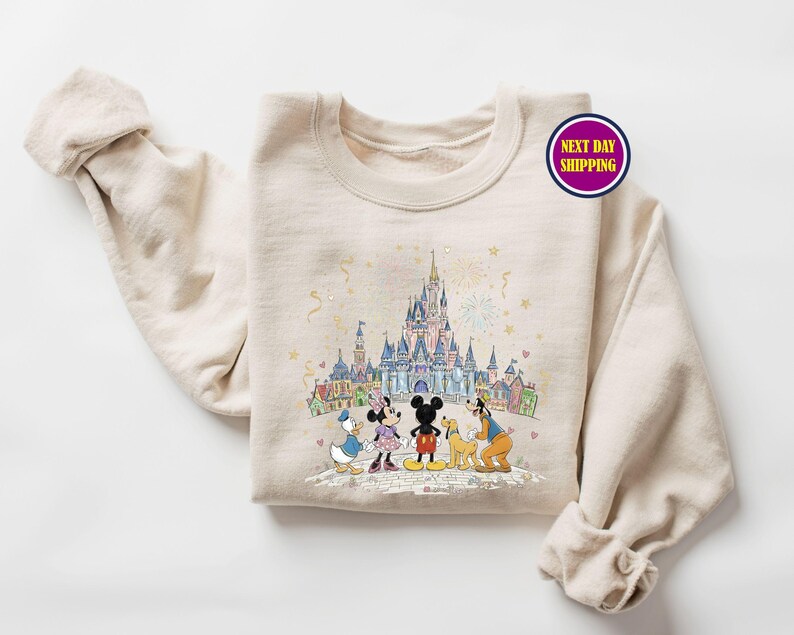 May include: Beige crewneck sweatshirt featuring a colorful illustration of a castle and cartoon characters. The graphic includes Mickey Mouse, Minnie Mouse, Donald Duck, and Goofy. The sweatshirt has the text "Next Day Shipping" on a circular sticker.
