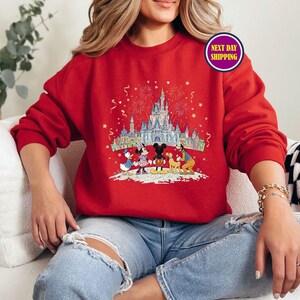 May include: Red sweatshirt featuring a colorful graphic of a castle, fireworks, and Disney characters. The design includes Mickey Mouse, Minnie Mouse, Donald Duck, and Goofy. The sweatshirt is a solid red color.