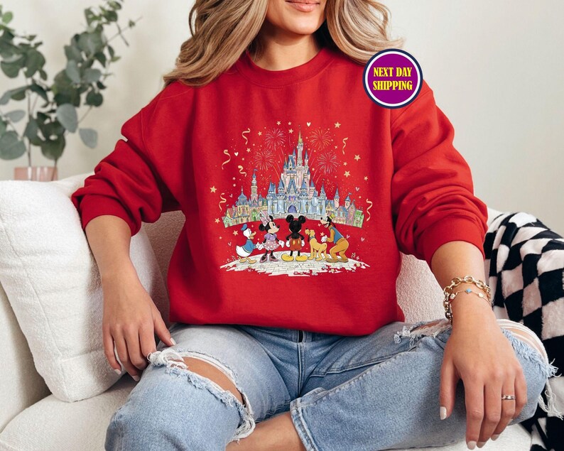 May include: Red sweatshirt featuring a colorful graphic of a castle, fireworks, and Disney characters. The design includes Mickey Mouse, Minnie Mouse, Donald Duck, and Goofy. The sweatshirt is a solid red color.