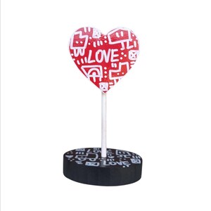 Peut inclure: Une sucette en forme de cœur rouge avec des motifs de style graffiti blancs et le mot "LOVE" dessus. La sucette est sur un support noir avec un design similaire.