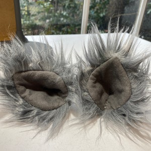 May include: A pair of gray faux fur wolf ears with a dark gray interior. The ears are fluffy and have a realistic shape, perfect for cosplay or costume use.