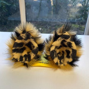 May include: A pair of faux fur cat ears with a leopard print design. The ears are tan with black spots and are surrounded by a ring of tan and black fur. The ears are attached to a yellow headband.