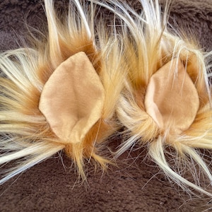 May include: A pair of lion-themed costume ears. The ears are made of tan fabric and surrounded by a mix of brown, tan, and white faux fur. The ears are designed to be worn as part of a costume or cosplay.