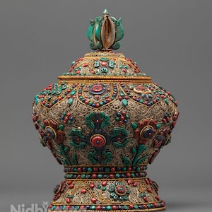 May include: An elaborate, lidded vessel with detailed filigree and vibrant stone inlays. The vessel has a gold-toned metal base and lid, embellished with red, blue, and green stones. A turquoise finial crowns the lid, providing a striking contrast to the design.