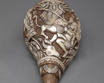 May include: A conch shell featuring intricate silver-toned metal relief carvings of figures and animals on a textured brown surface. The detailed metalwork covers the exterior of the shell, which displays the word NIDHIRATNA at the base.