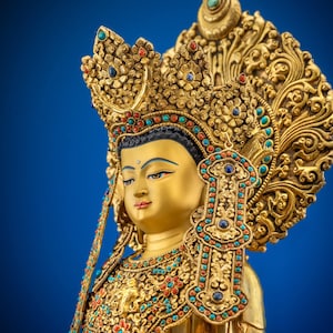 Peut inclure: Une statue dorée d'une figure sereine, ornée d'une coiffe et de bijoux élaborés, ornés de pierres précieuses colorées. La statue a une expression calme sur un fond bleu vif. Le texte "Nidhiratna" est visible en bas.