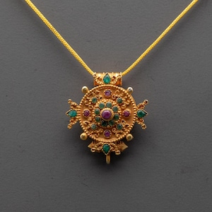 May include: A gold-colored pendant necklace with a circular design. The pendant features a central cluster of small, round, purple gemstones surrounded by green and purple gemstones. The necklace chain is a thin, gold-colored chain.