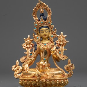 May include: A gold-colored statue of a seated deity with intricate details. The statue features a crown, turquoise accents, and ornate floral designs. The deity is seated on a lotus base, with the text "Nidhiratna" visible at the bottom.