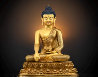 May include: A gold-colored Buddha statue with a black hair bun and a serene expression. The statue is seated in a meditative pose with hands in the Dhyana mudra. The statue is on a lotus-shaped base.