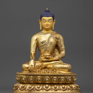 Könnte beinhalten: Eine goldene Buddha-Statue in meditativer Sitzhaltung. Die Statue hat eine blaue Ushnisha, detaillierte Kleidung und einen ruhigen Gesichtsausdruck. Der Sockel ist aufwendig mit floralen Mustern gestaltet. Die Statue ist eine Darstellung buddhistischer Kunst.