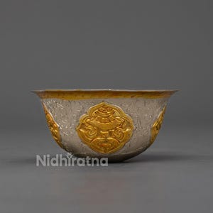 May include: A silver bowl with gold accents and ornate detailing. The bowl features a gold rim and gold decorative elements. The brand name "Nidhiratna" is visible on the side of the bowl. The bowl is likely used for decorative or ceremonial purposes.