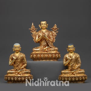 Puede incluir: Tres estatuas doradas de Buda en posición sentada. Las estatuas están sobre bases ornamentadas y tienen túnicas detalladas. La estatua central tiene un elemento decorativo detrás. Las estatuas están sobre un fondo gris. El texto "Nidhiratna" está en la parte inferior.