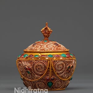 Può includere: Scatola decorativa dorata e ornata con coperchio a cupola. La scatola presenta intricati motivi a filigrana ed è adornata con pietre preziose rosse e verdi. Un piccolo terminale decorativo si trova sul coperchio. Il testo "Nidhiratna" è visibile.