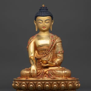 Peut inclure: Statue dorée de Bouddha en position de méditation, tenant un petit bol. La statue a des cheveux bleu foncé et des ornements rouges et dorés. La base est ornée d'un motif de lotus. Le texte "Nidhiratna" est en bas.