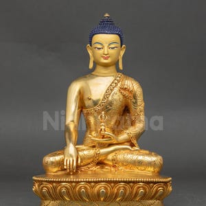 May include: A gold Buddha statue in a seated pose, with detailed patterns on the robes and base. The statue features blue hair and a calm expression. The statue is set on a lotus flower base.