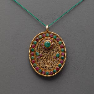 May include: An oval gold pendant featuring intricate filigree metalwork in a floral pattern, centered with a single round green stone. The border is set with alternating round red and green stones, suspended from a thin green cord necklace.