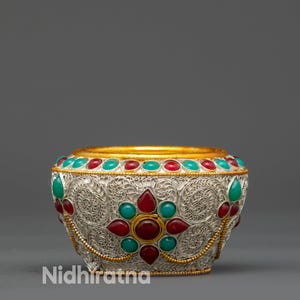 May include: Ornate silver bowl with gold trim and inlaid gemstones. The bowl features a floral design with red and turquoise stones. The text "Nidhiratna" is visible on the lower left side of the bowl.