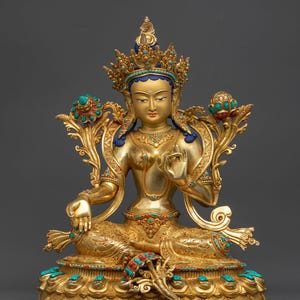 Könnte beinhalten: Eine goldene Statue einer sitzenden Gottheit, geschmückt mit einer juwelenbesetzten Krone und aufwendigen Details. Die Figur hat einen ruhigen Ausdruck und ist mit türkisen und roten Akzenten verziert. Die Statue ist eine Darstellung einer spirituellen Figur.