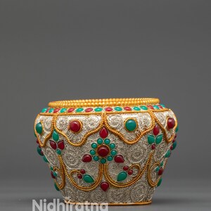 May include: Silver-colored decorative bowl with intricate gold filigree and inlaid red and green gemstones. The bowl features a textured surface and a gold rim. The text "Nidhiratna" is visible on the side.