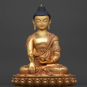 Peut inclure: Statue dorée de Bouddha en position assise, tenant un petit bol. La statue a des cheveux bleu foncé et est ornée d'accents rouges et turquoise. La base est ornée d'un motif de lotus. Le texte "Nidhiratna" est visible en bas.