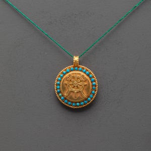 Puede incluir: Colgante dorado con borde turquesa y diseño de rueda. El colgante cuelga de un cordón verde. El diseño de la rueda está en el centro del colgante.