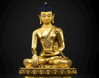 May include: A golden Buddha statue in a seated position, with a blue head covering. The statue is on a decorative base and holds a small bowl. The image is set against a dark background. The statue is from Nidhiratna.