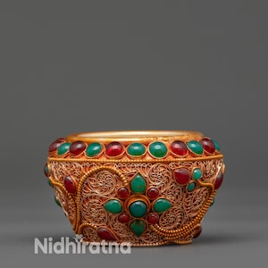 May include: Ornate gold-colored bowl with red and green gemstones. The bowl features intricate filigree patterns and is adorned with a row of gemstones around the rim. The text "Nidhiratna" is visible on the side.